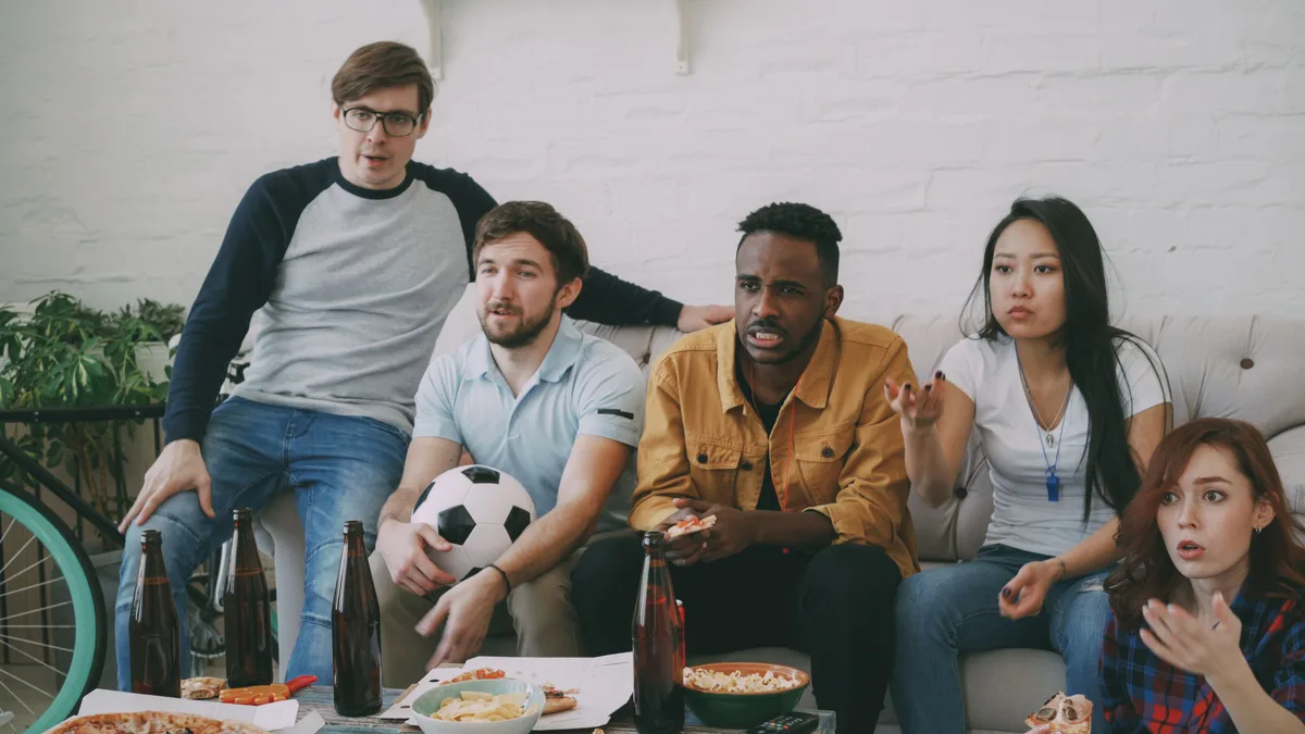 Cinco amigos reunidos na sala de casa assistindo a um jogo de futebol na televisão