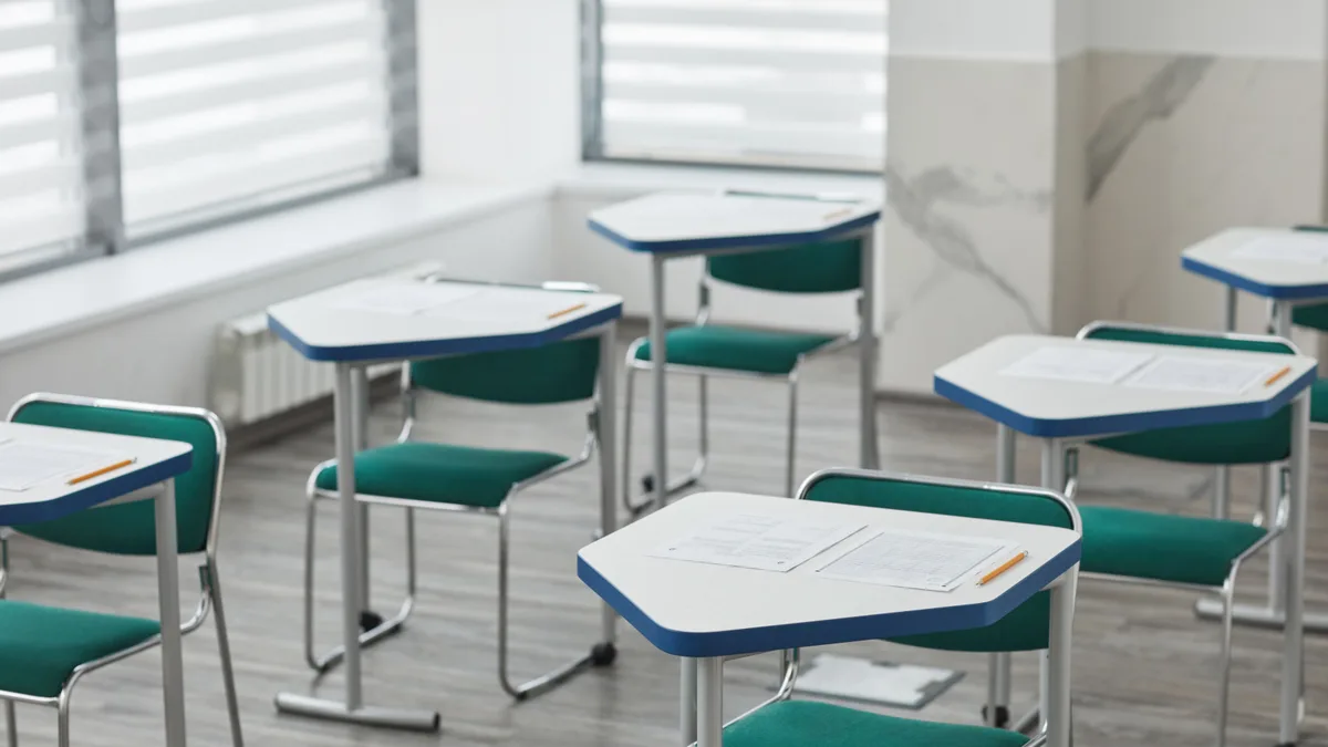 Sala de aula vazia com carteiras enfileiradas, cada uma com folhas de prova e lápis, pronta para o dia do exame