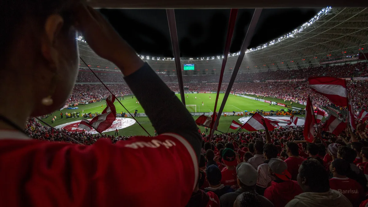 Estádio brasileiro cheio durante partida de futebol à noite, com torcida animada