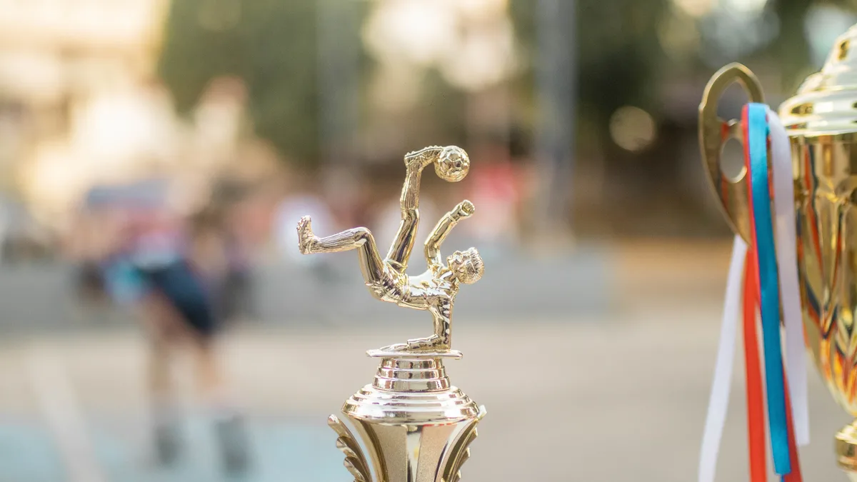 Troféu dourado de futebol com a figura de um jogador em bicicleta, ao lado de outra taça com fitas