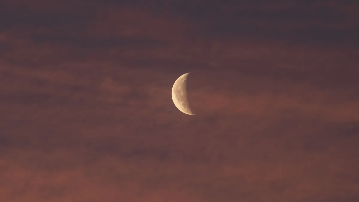 Lua crescente vista entre nuvens finas, em céu de tom avermelhado no fim do entardecer