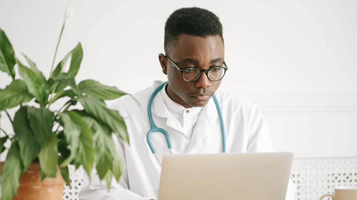 Médico de jaleco branco consulta um laptop em sala clara, com planta ao lado