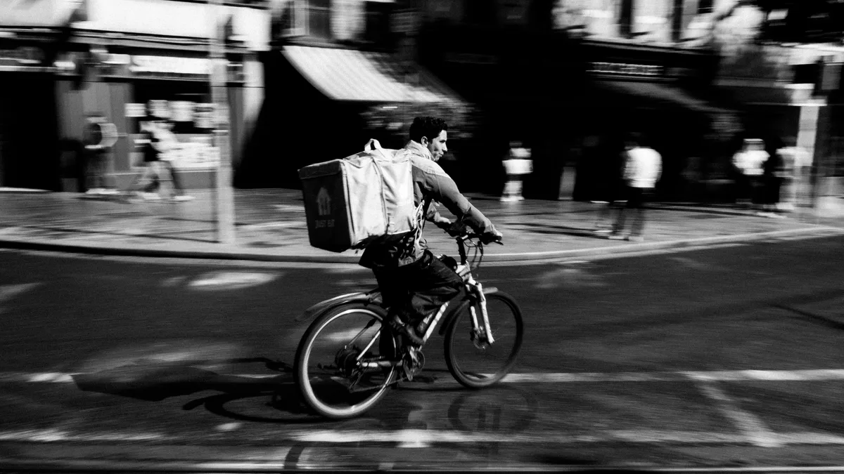 Entregador de bicicleta com mochila térmica atravessa rua movimentada da cidade