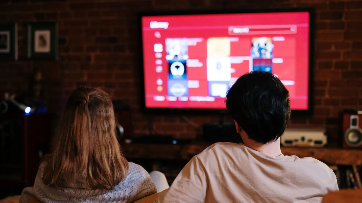 Casal sentado no sofá assistindo a uma série na televisão, em sala com parede de tijolos