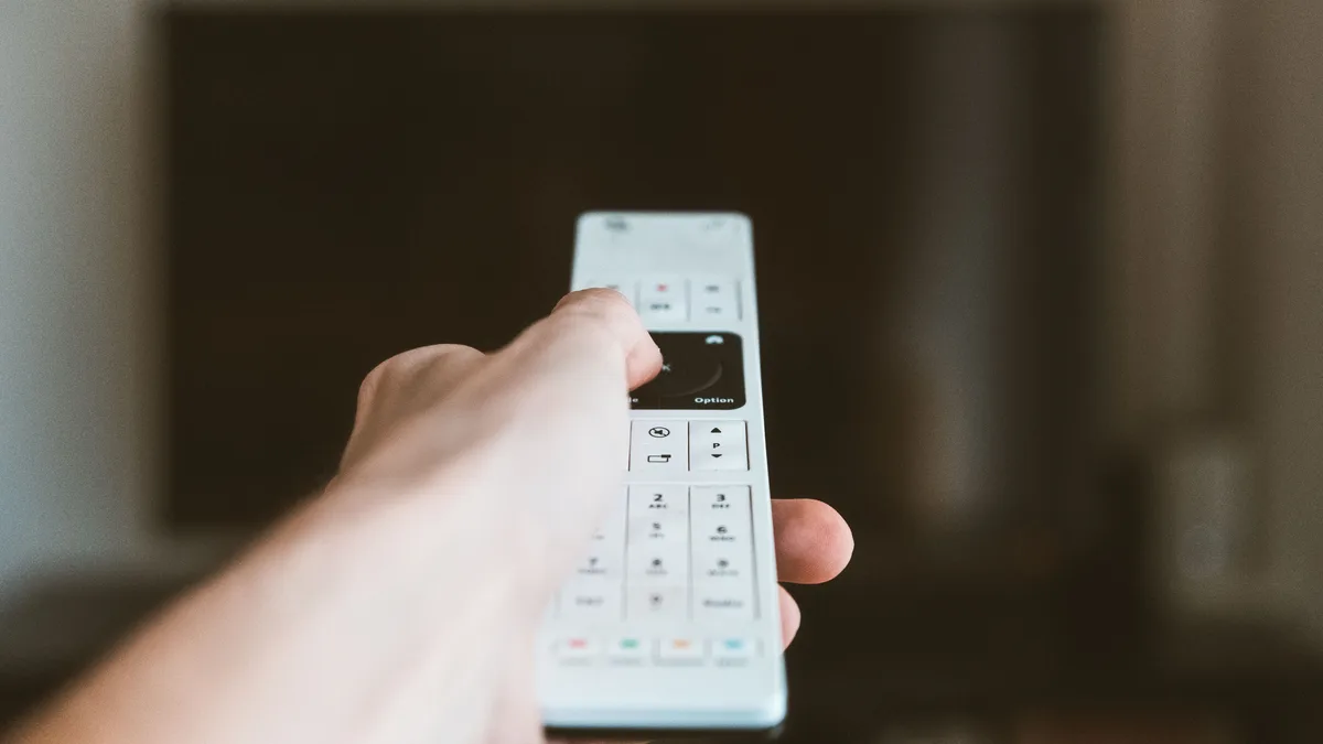 Mão segurando um controle remoto em frente a uma televisão, dentro de casa