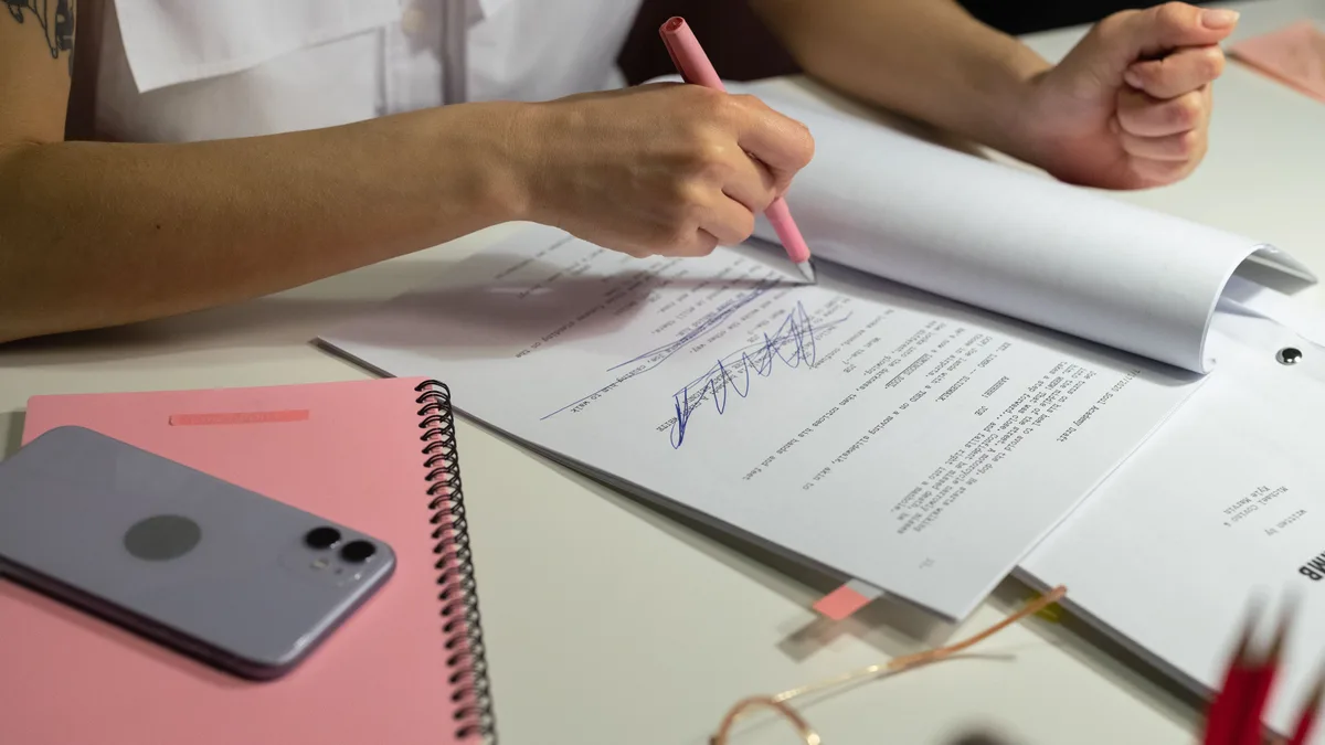 Mãos assinando um documento sobre a mesa, ao lado de caderno e caneta