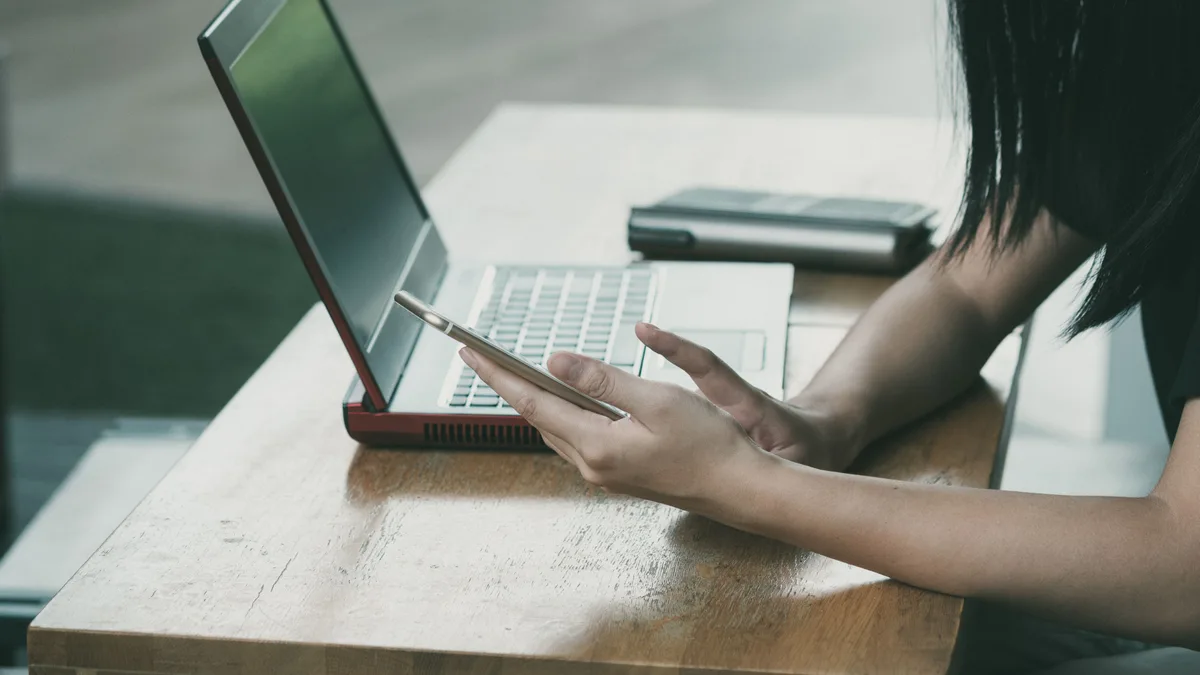 Jovem consulta o celular diante do notebook em uma mesa