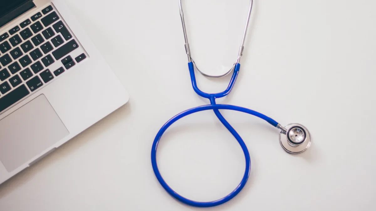 Estetoscópio azul enrolado ao lado de um notebook, sobre mesa branca de estudo