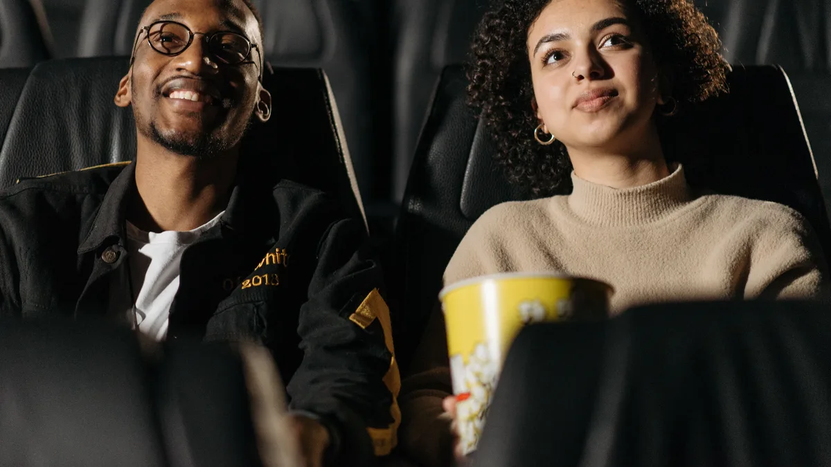 Casal jovem sorrindo em sala de cinema, dividindo um balde de pipoca