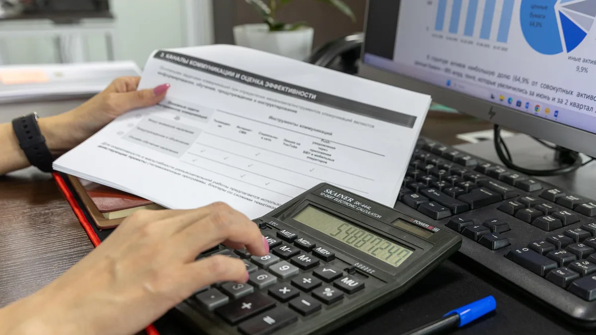 Pessoa calcula dados financeiros com calculadora e documentos, em mesa de escritório