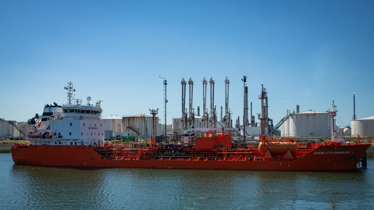 Navio-tanque vermelho atracado em porto industrial, com tanques de armazenamento ao fundo